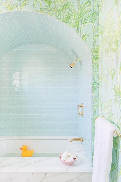 sky blue glass tiled barrel ceiling, a marble-clad drop-in bathtub is fixed under an antique brass shower kit fixed against sky blue mini subway wall tiles, while walls are clad in green and blue wallpaper.