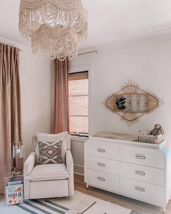 Welcoming nursery is illuminated by a beaded chandelier and features a modern white curved dresser topped with a changing pad and positioned under a rattan mirror. An acrylic accent table accented with a nickel and glass lamp is placed beside an off-white glider, while windows are covered in brown linen curtains.