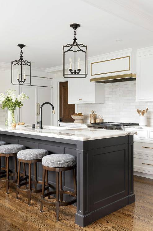 Backless stools sit at a black center island topped with a statuary marble countertop holding a sink with a matte black gooseneck faucet lit by two black gothic lanterns.