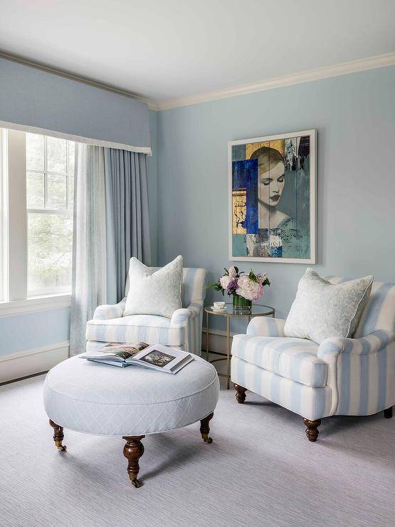 Blue bedroom sitting area is furnished with two blue stripe French chairs placed on a gray rug on either side of a round brass and glass accent table positioned beneath an art piece hung from a blue wall. The chairs are matched with a round blue lattice ottoman and complemented with a window covered in blue curtains hung beneath a blue valance.