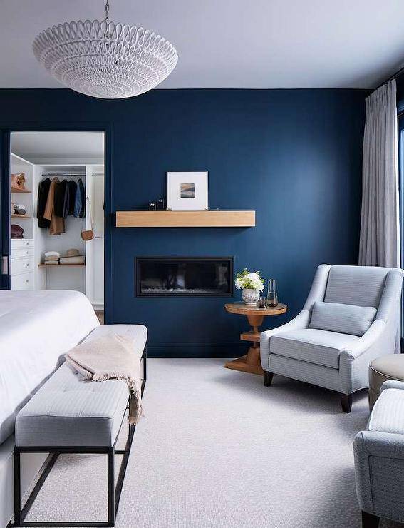 A modern inset fireplace with a styled chunky mantel is positioned in a blue painted bedroom wall beside a glass top round wooden accent table matched with a gray striped upholstered chair.
