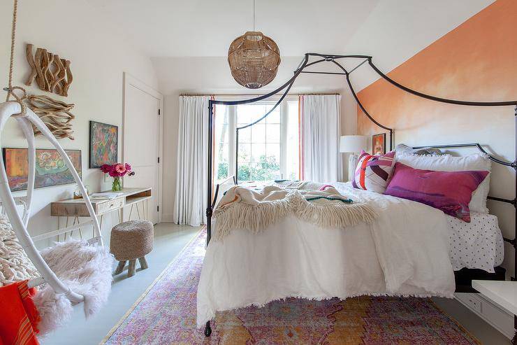 This stylish, boho chic kid's bedroom boasts an orange ombre accent wall positioned behind an iron canopy bed. The bed sits on a pink a pink and orange vintage wool rug and is accented with pink and purple pillows, while a white hanging chair is fixed in a corner.