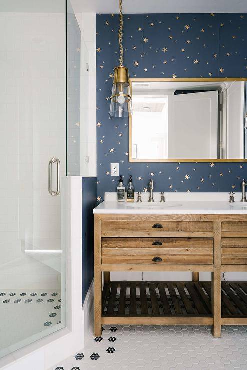 Kid's bathroom features a brass mirror mounted on blue and gold starburst wallpaper over a reclaimed wood dual washstand lit by a glass and brass sconce and a corner walk in shower.