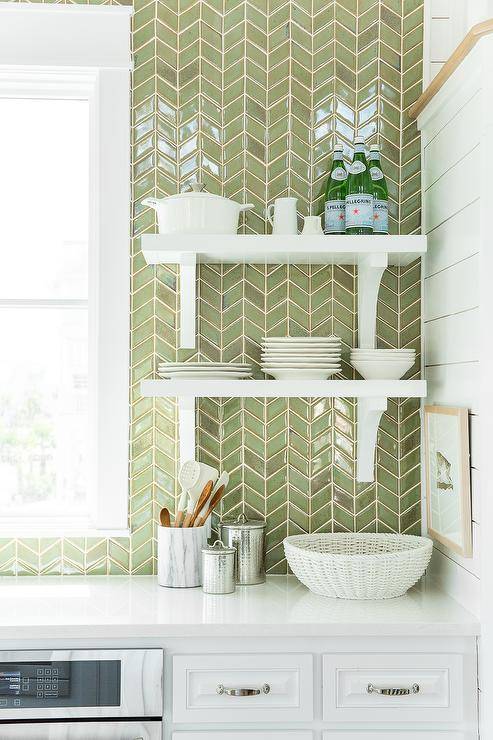 White and green kitchen features white cabinets paired with white quartz countertops and a ceiling height green herringbone tiled backsplash lined with stacked white shelves with corbels.