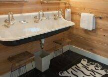 Chic cottage kid's bathroom features black floor tiles covered in black and white zebra print bath mat positioned in front of a black and white trough sink finished with three satin nickel faucets and mounted to a wood paneled wall above two iron and wood step stools. The sink is fixed below two curved vanity mirrors with shelves illuminated by nickel sconces.