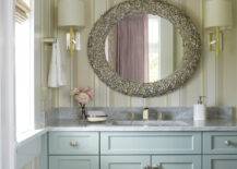 Coastal bathroom features Laura Ashley Eaton Stripe Wallpaper framing Made Goods Jules Mirror flanked by Barbara Barry Refined Rib Sconces over blue vanity painted Sherwin Williams Quietude topped with White Carrera Extra Marble.