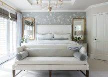 Gold leaf mirrors over white French nightstands in a chic white and gray bedroom finished with crystal lamps. A white settee with gray bolster pillows is finished at the foot of the bed with a sophisticated touch.