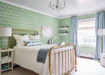 green wallpaper features a brass vintage bed topped with a gray fringe throw blanket and a white and blue lumbar pillow tall white nightstand lit by a glass and brass lamp and a white desk placed beneath a Greek key pin board