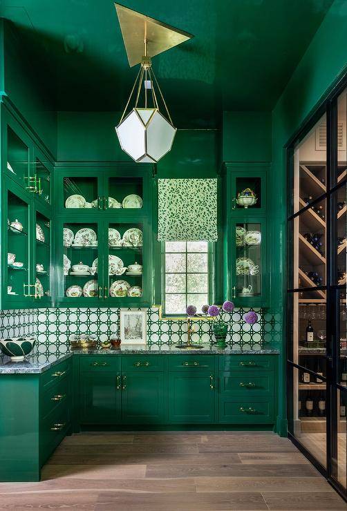 Emerald green pantry is lit by a modern brass chandelier fixed to a glossy emerald green ceiling and lighting emerald green lacquer cabinets with lucite pulls. The cabinets are topped with a gray marble countertop fixed against white and green lattice backsplash tiles under stacked glass front upper cabinets. A white and green roman shade hangs from a small window above a round brass sink.