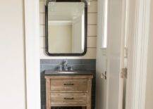Small white and gray cottage bathroom showcases an eye catching rustic oak single washstand finished with a slated wood shelf, oil rubbed bronze hardware, and a soapstone countertop fitted with an oval undermount sink paired with a polished nickel vintage cross handle faucet. The faucet is positioned in front of gray linear backsplash tiles and beneath a curved black vanity mirror mounted on a white shiplap wall lit by two glass light pendants.