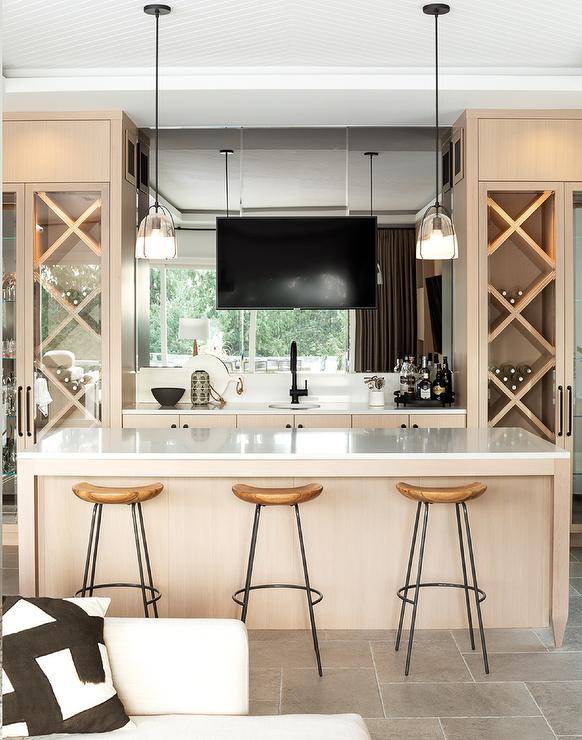 Three backless wood and iron stools are placed at a tan bar island. Behind the island, a TV is mounted in front of a mirrored backsplash over a small round sink with a matte black faucet. The TV is flanked by glass front built-in wine rack cabinets.
