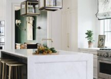 White, gold, and black kitchen is lit by gold and black French lanterns hung over a marble waterfall island top finished with a sink and matched with black and gold backless counter stools.