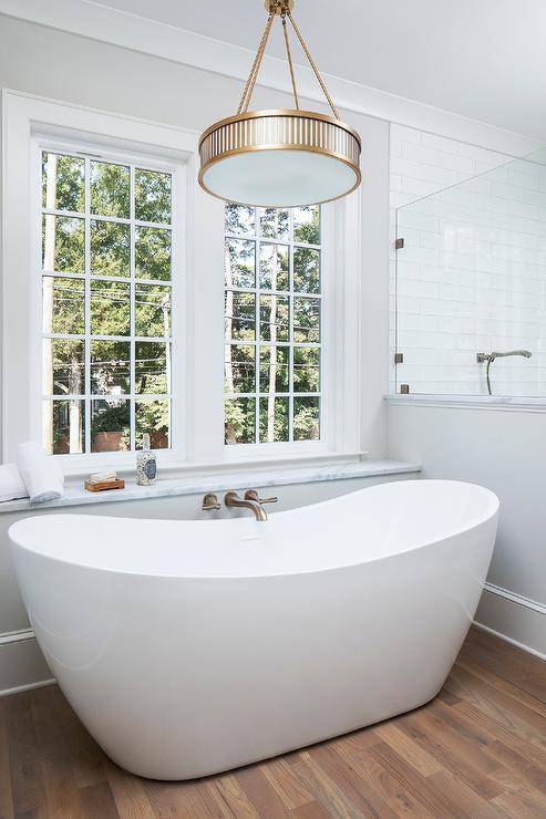 Brushed gold wall mount tub filler designed with an oval freestanding tub illuminated with a brass hanging light in a transitional bathroom design. A window over the tub adds lovely natural light to an already bright and airy bathroom design.