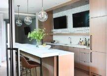 Large modern wet bar design features side-by-side TVs mounted on gray marble chevron backsplash tiles over light brown veneer cabinets. Brown leather stools sit at a quartz topped wet bar under clear globe pendants.