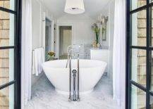 White cottage bathroom features a white tapered drum pendant light which hangs over a freestanding bathtub placed in front of steel doors dressed in white curtains which open to the deck.