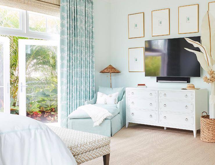 Art surrounds a television placed on a white Greek key dresser in a bedroom boasting a blue chaise lounge positioned in a corner in front of glass doors covered in blue curtains layered over bamboo roman shades.