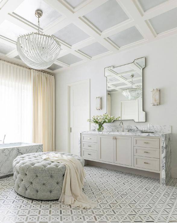 An art deco chandelier hangs from a gray coffered ceiling over a gray tufted ottoman placed in an elegant bathroom on marble parquet-style floor tiles. An art deco vanity mirror is mounted above a light gray washstand finished with a white and gray marble waterfall countertop lit by marble sconces. Ivory curtains hang from a window located over a marble rectangular bathtub.