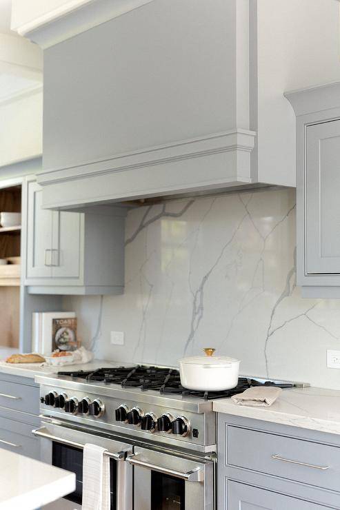 Kitchen features a light blue wooden kitchen hood over a stainless steel dual range on a gray and white marble look slab backsplash and blue cabinets accented with nickel pulls.