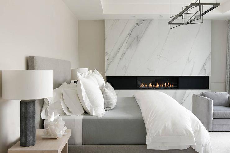 Gray modern bedroom boasts a modern gray bed accented with white and gray bedding and flanked by tan wood nightstands topped with black lamps. The room is warmed by a lava rock fireplace fixed facing gray velvet accent chairs