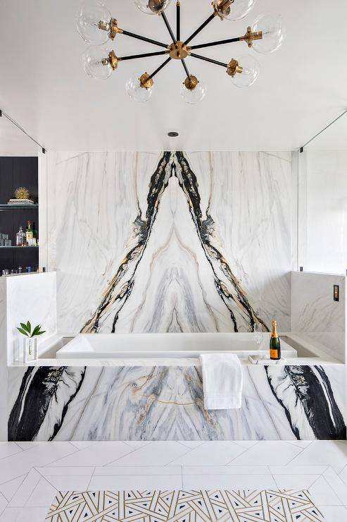 Master bath features gold, black and gray bookmatched marble on a wall placed over a matching marble clad bathtub illuminated by an atom chandelier.