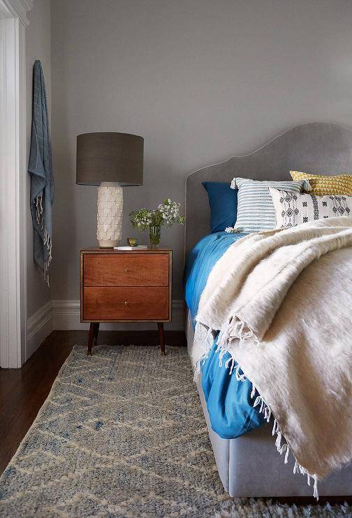 A gray velvet upholstered bed dressed in bold blue bedding stands next to a wood mid-century modern nightstand and an ivory lamp alongside a blue wool rug.