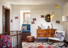 Vintage nursery features a burl wood dresser with pink tassel pulls, an oval wooden crib with colorful blanket, a white vintage nursery rocker accented with an Otomi pillow illuminated by a brass floor lamp, a mini window seat with blue cushion and a colorful rug.