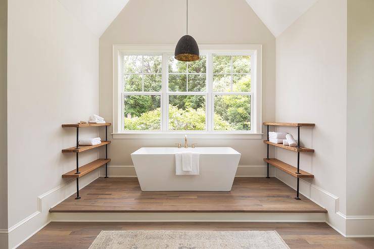 Industrial pipe and salvaged wood shelves create a unique feature flanking a rectangular freestanding tub completed with a brass faucet under a window and vaulted ceiling displaying a black hanging drum pendant. A step up feature adds to an appealing surface for the freestanding tub with a beige runner in front of the step.