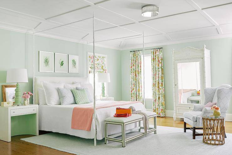 Pastel-colored bedroom is accented with spring green walls and a white coffered ceiling. Three art pieces hang over a white canopy bed finished with a white upholstered headboard and white bedding accented with a peach-colored throw blanket. The bed is flanked by white nighstands topped with green lamps complementing a green bench placed at the foot of the bed. A window dressed in floral curtains is located beside a white bamboo armoire completed with a mirror.