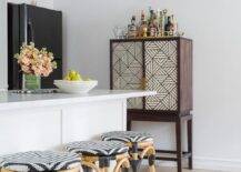 A tall wooden bar cabinet with cream and brown geometric doors sits to the side of a white kitchen peninsula seating backless rattan stools lit by a round brass flush mount.