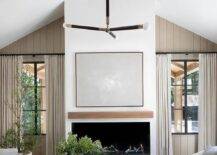 A light gray abstract art piece hangs over a modern fireplace fitted with a wood floating mantel and flanked by windows framed by taupe plank trim and covered in beige curtains.