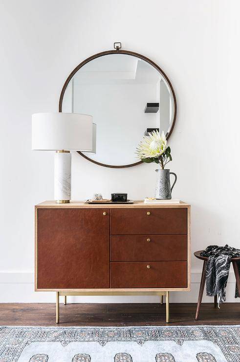 Perfectly aligned, this mid-century modern cabinet boasts a warm wood finish with a brass outline and legs. West Elm Marble Pillar Table Lamp is displayed atop the cabinet along with a tray, accessories, and vase with flowers. Large round iron mirror hangs over the cabinet on a white wall highlighting an eclectic foyer design while a small round wood stool against oversized white baseboards is draped with a black and white throw. Brown oak flooring is layered with a blue and gray patterned rug creating a cozy, homey touch to a well-designed foyer.