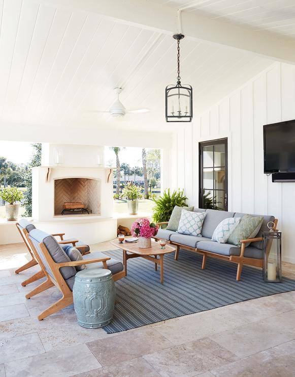 Teak mid-century modern outdoor sofa and chairs designed facing an oval slatted teak coffee table atop a blue striped outdoor rug. This lovely covered patio boasts a white modern ceiling fan from a vaulted plank ceiling finished with a white outdoor fireplace.