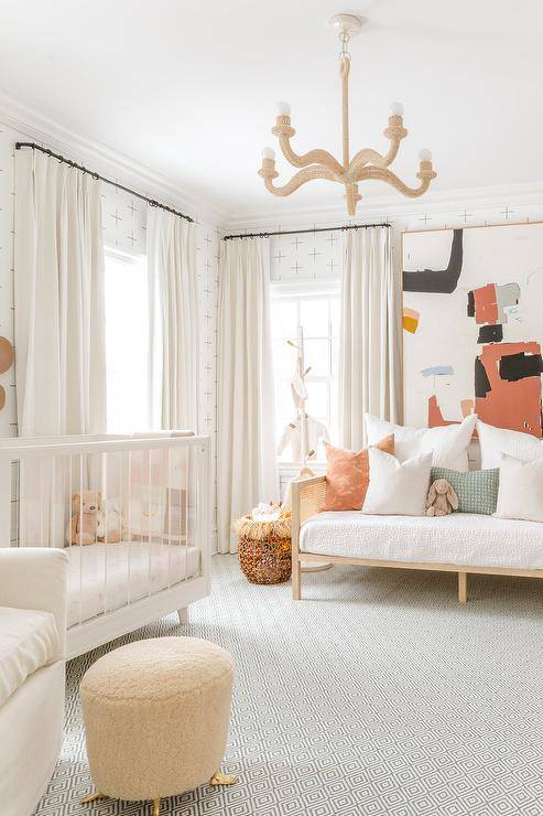 Charming gender-neutral nursery is lit by a rope chandelier hung over a gray diamond print rug, while walls are clad in minimalistic white and black wallpaper. A window dressed in white curtains is located next to a white wood and acrylic crib, as a large abstract art piece hangs over a daybed.