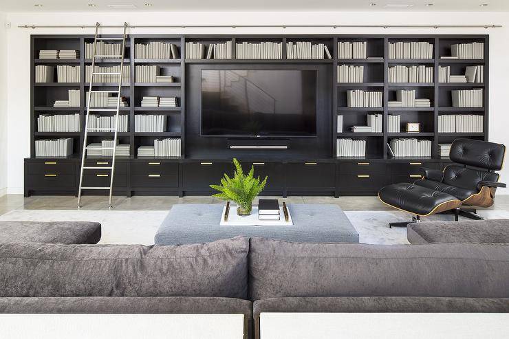 A gray u-shaped sectional sits on a light gray rug in a modern living room furnished with a blue tufted bench and an Eames Lounge Chair & Ottoman placed in front of a floor-to-ceiling black built-in tv unit fitted with a ladder on rails.