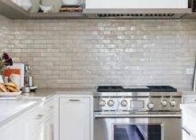 White kitchen features taupe glazed brick backsplash tiles with gray floating shelves, a wolf range under a white hood and white cabinets with nickel pulls.