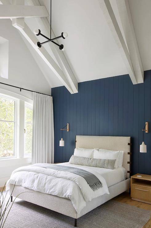 Positioned against a vertical blue plank wall, a boucle and leather bed is topped with a long gray lumbar pillow and a gray fringe throw blanket. The bed sits on a gray rug between light brown wooden nightstands lit by wall sconces.
