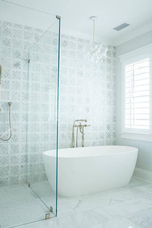 An oval freestanding bathtub lit by a glass bubbles chandelier sits on marble floor tiles and is paired with a polished nickel floor mount tub filler mounted in front of a gray marble lattice tiled wall. The tub is positioned beside a frameless glass shower.