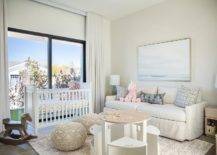 Welcoming nursery is styled with macrame poufs placed on a gray rug at a round two-tone play table with white chairs. n ocean art piece hangs over an off-white couch topped with gray and purple pillows lit by turquoise blue cork lamps. A white crib sits in front of sliding glass doors covered in white curtains.