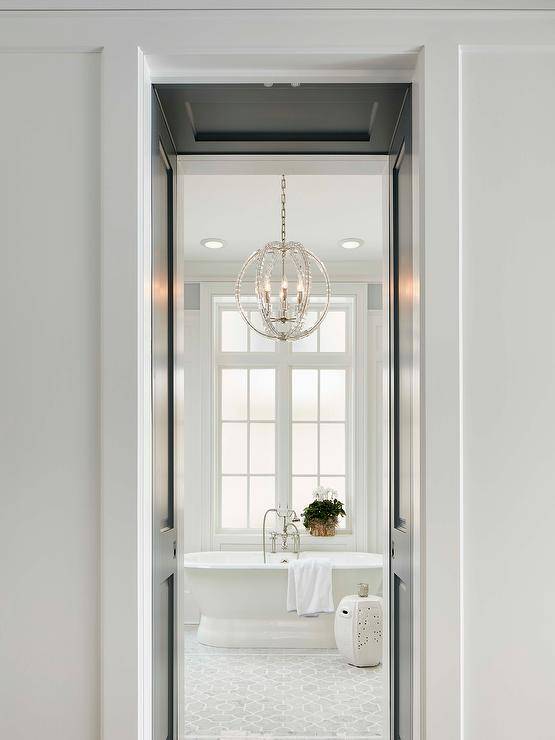 A doorway opens to a bathroom filled with a clear beaded sphere chandelier illuminating a roll top tub atop a gray mosaic marble tiled floor.