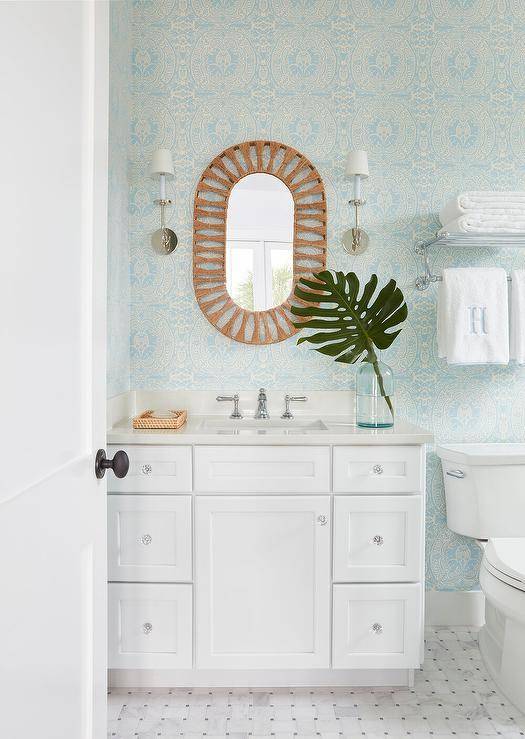 Nickel scones hang on either side of an oval rope mirror hung from a powder room wall clad in blue damask wallpaper over a white washstand adorned with glass knobs and polished nickel faucet kit. A white porcelain toilet is mounted against marble basketweave floor tiles under a towel rack.