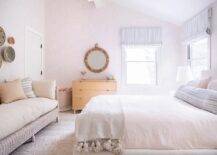 Decorative baskets hang from a light pink wallpapered bedroom wall over a white wicker sofa with beige cushions and earth tone pillows, as a round rope mirror is mounted above a beige wooden dresser. Light pink pillows are layered behind a large blue striped lumbar pillow.