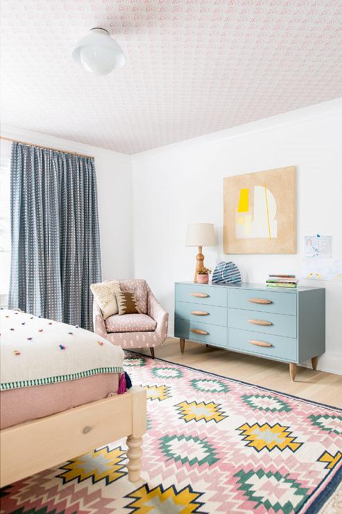 Stylish eclectic girl's bedroom tan and yellow abstract art piece hung beneath a ceiling clad in pink wallpaper and over a gray dresser lit by a wooden lamp. A pink chair sits in a corner on a pink, green, blue, and yellow rug and in front of a window dressed in pink and blue curtains.