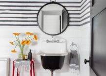 Farmhouse styled powder room with shiplap walls, small trough sink, and a barn red stool accent table. Black and white striped wallpaper adds a stylish dimension to the bathroom above the shiplap trim displaying a barn red vintage barn sconce above a round black vanity mirror.