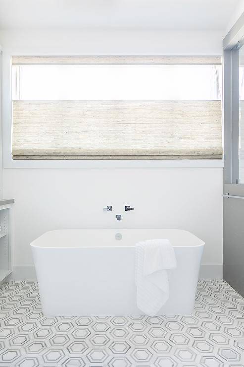 A chrome tub filler is mounted beneath a window and over a rectangular freestanding bathtub placed on white and gray marble hexagon floor tiles.