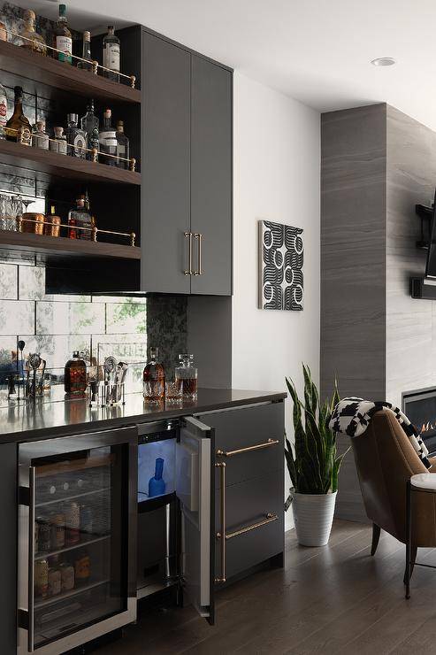 Modern living room wet bar features a mini ice maker, a mini beverage fridge, large antiqued mirrored subway tiles, black flat front cabinets and wood and brass shelves.