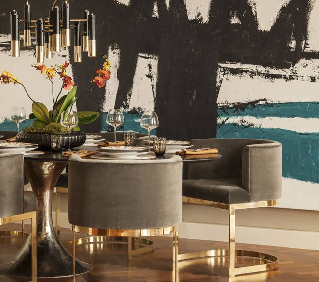 Chic dining room features large teal and black abstract art framing black and gold art deco chandelier over black and gold dining table surrounded by gray barrel back dining chairs over wood herringbone floor.