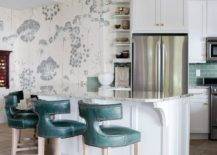 Teal leather t-back stools are placed at a granite countertop accenting a white kitchen peninsula wrapping around to built-in display shelves. A stainless steel double door refrigerator is recessed under white cabinets with brass knobs.