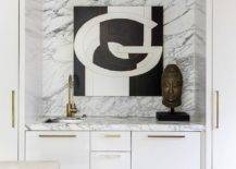 White flat-front wet bar cabinets accented with polished brass bulls and a marble countertop are built into a dining room alcove. The cabinets are finished with a sink and an aged brass gooseneck faucet mounted in front of a marble slab backsplash complemented with a black and white abstract art piece.
