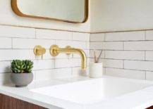 Lined with brass trim, white glazed subway wall tiles are fitted with a brass faucet kit mounted above an oak floating bath vanity and beneath a curved brass mirror.