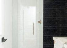 White and black mosaic floor tiles lead past a wall clad in black stacked tiles to a small walk-in shower boasting a frameless glass door and a brushed gold shower kit fixed to white subway staircase pattern surround tiles.